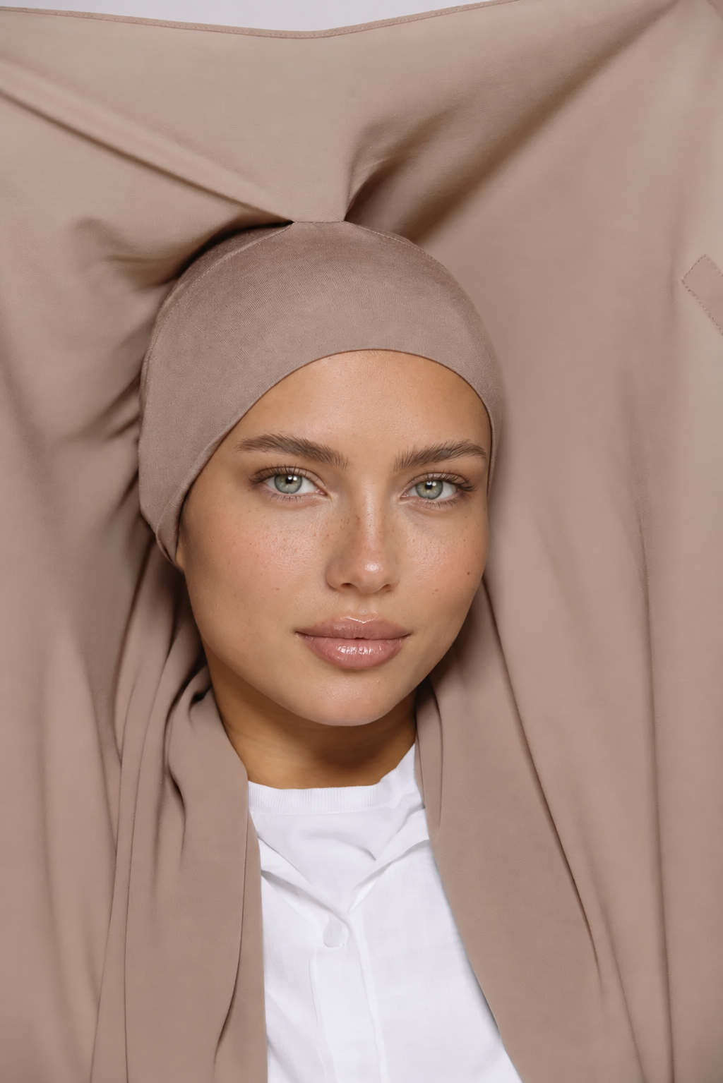 A woman wearing a soft taupe-beige hijab and matching head covering, styled with a white collared shirt. The fabric is draped elegantly behind her in a studio setting.