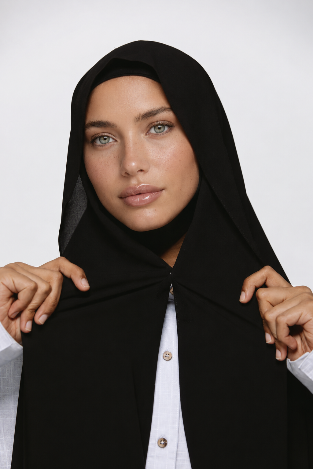A woman wearing a black hijab and a white collared shirt, holding the sides of the hijab with both hands against a plain light gray background.