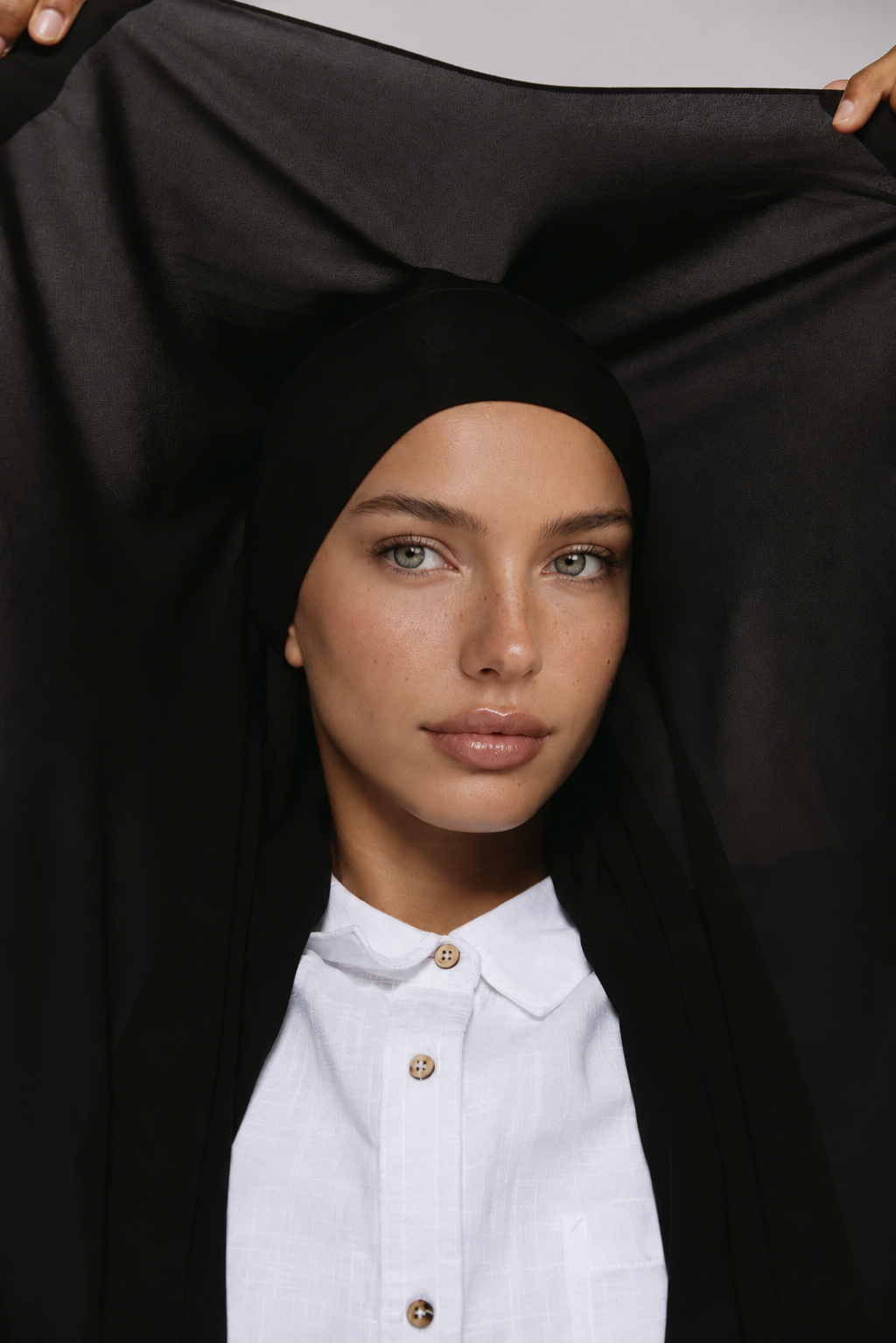 A woman wearing a black hijab and a white collared shirt, holding a large black fabric piece overhead to display its drape and coverage.