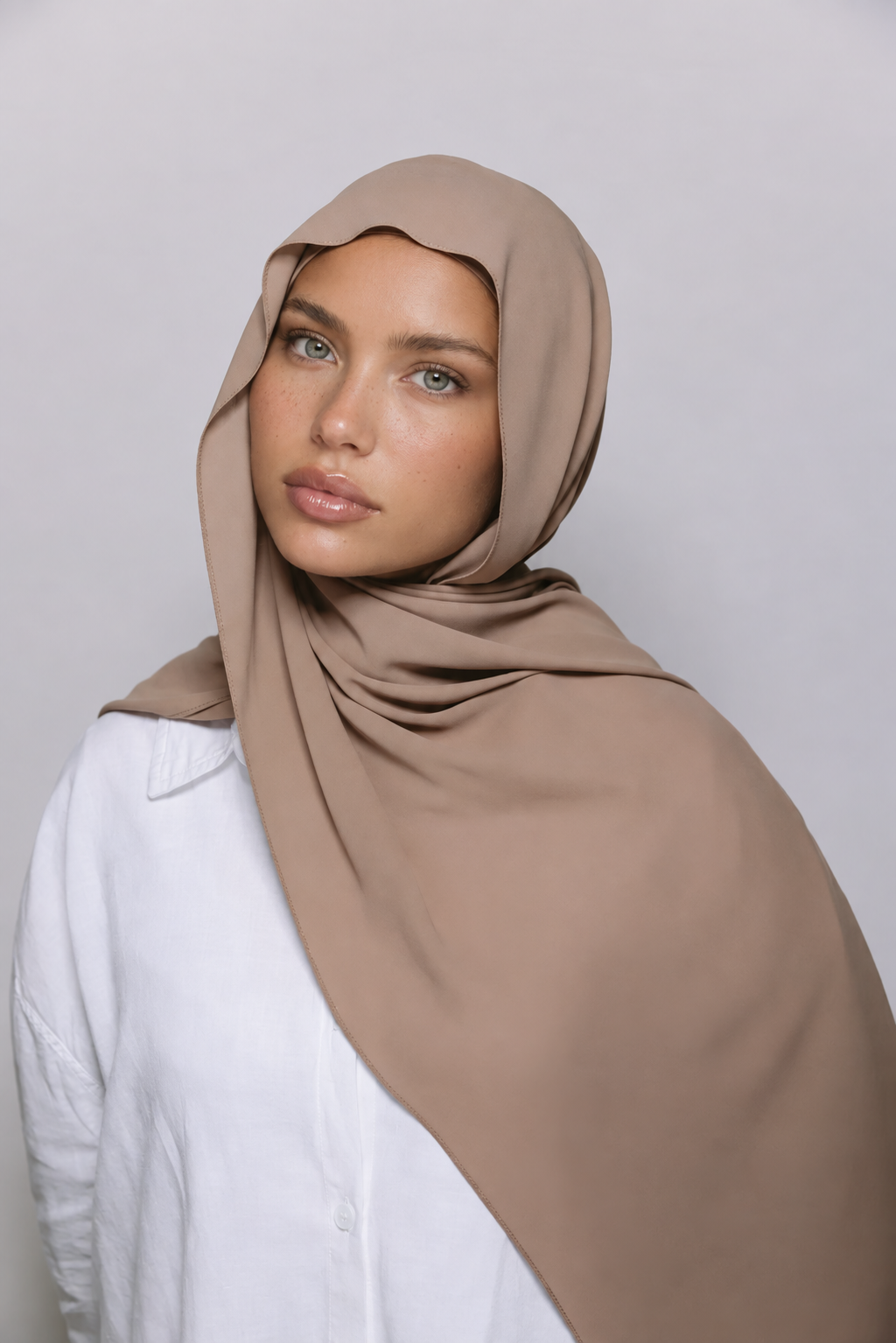 A woman wearing a solid taupe-beige hijab draped over her shoulders, paired with a white collared shirt, photographed against a neutral light gray background.