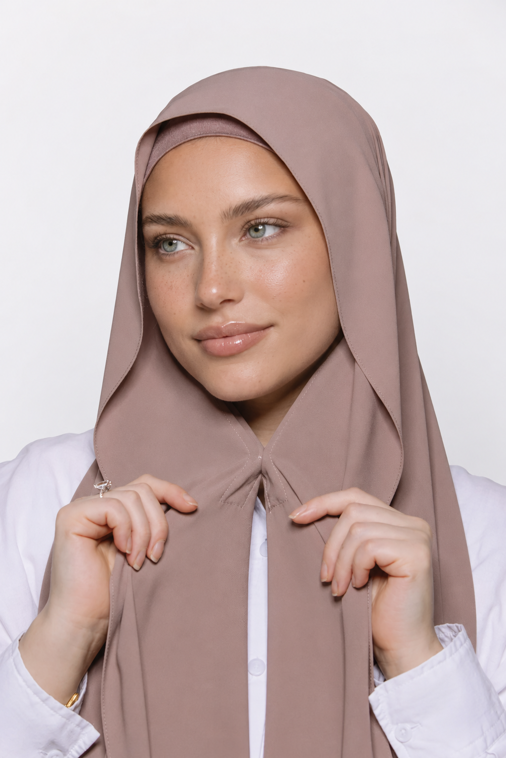 A woman wearing a draped dusty mauve hijab and a white collared shirt, posing against a clean white studio background.