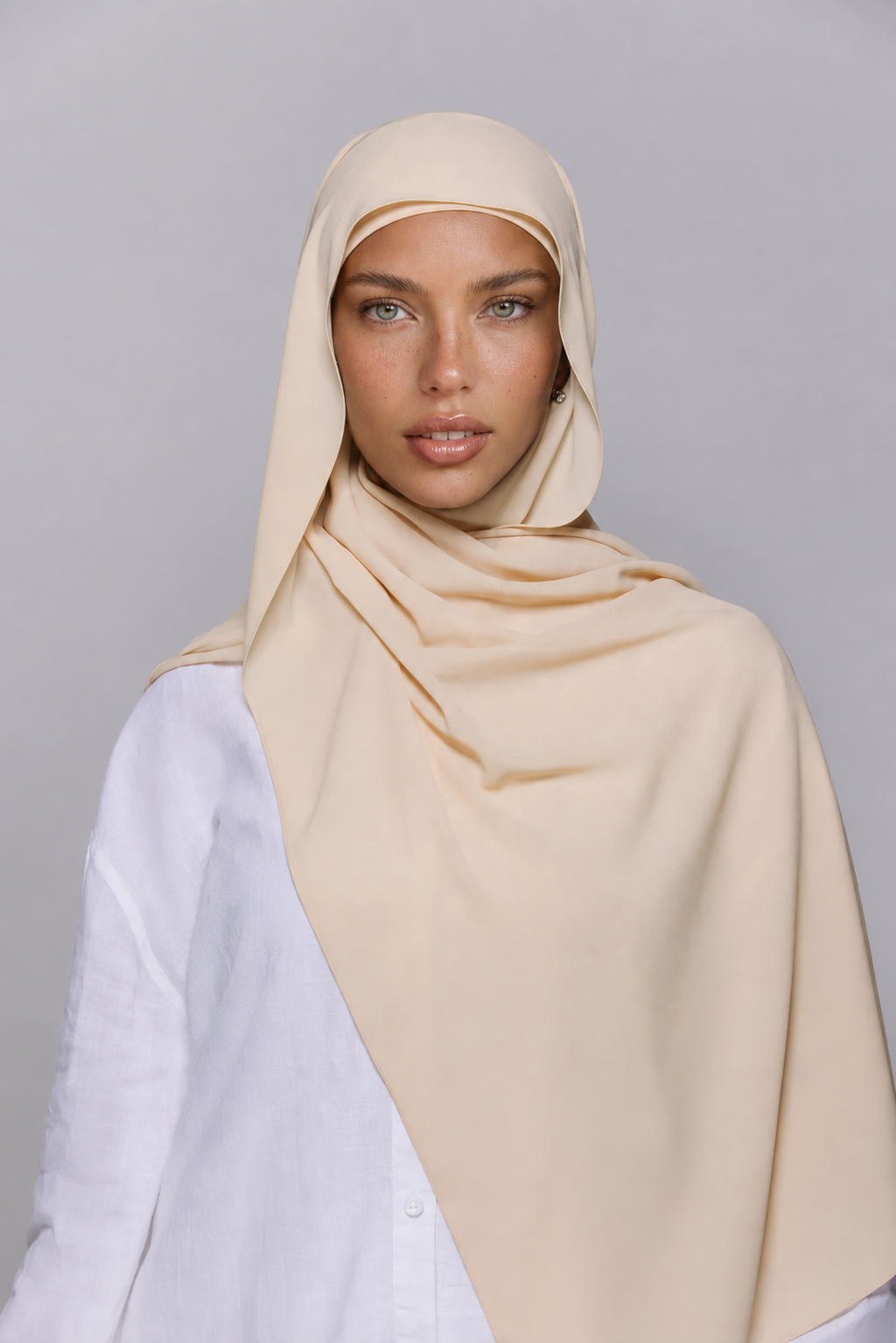 A woman wearing a light beige hijab and a white button-up shirt, photographed against a neutral gray studio background.