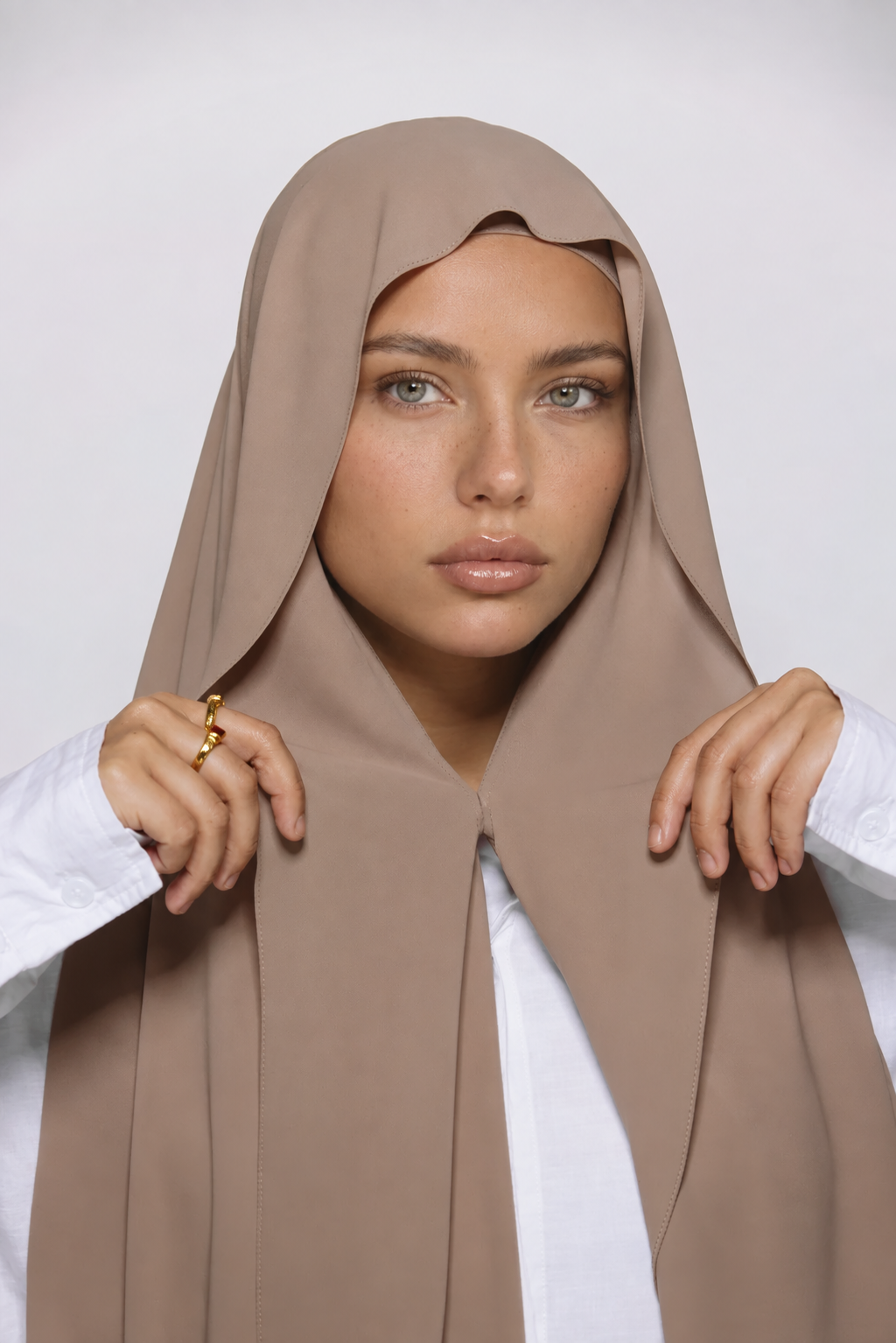 A woman wearing a taupe-beige hijab draped over her shoulders, styled over a white collared shirt, against a plain light gray studio background.