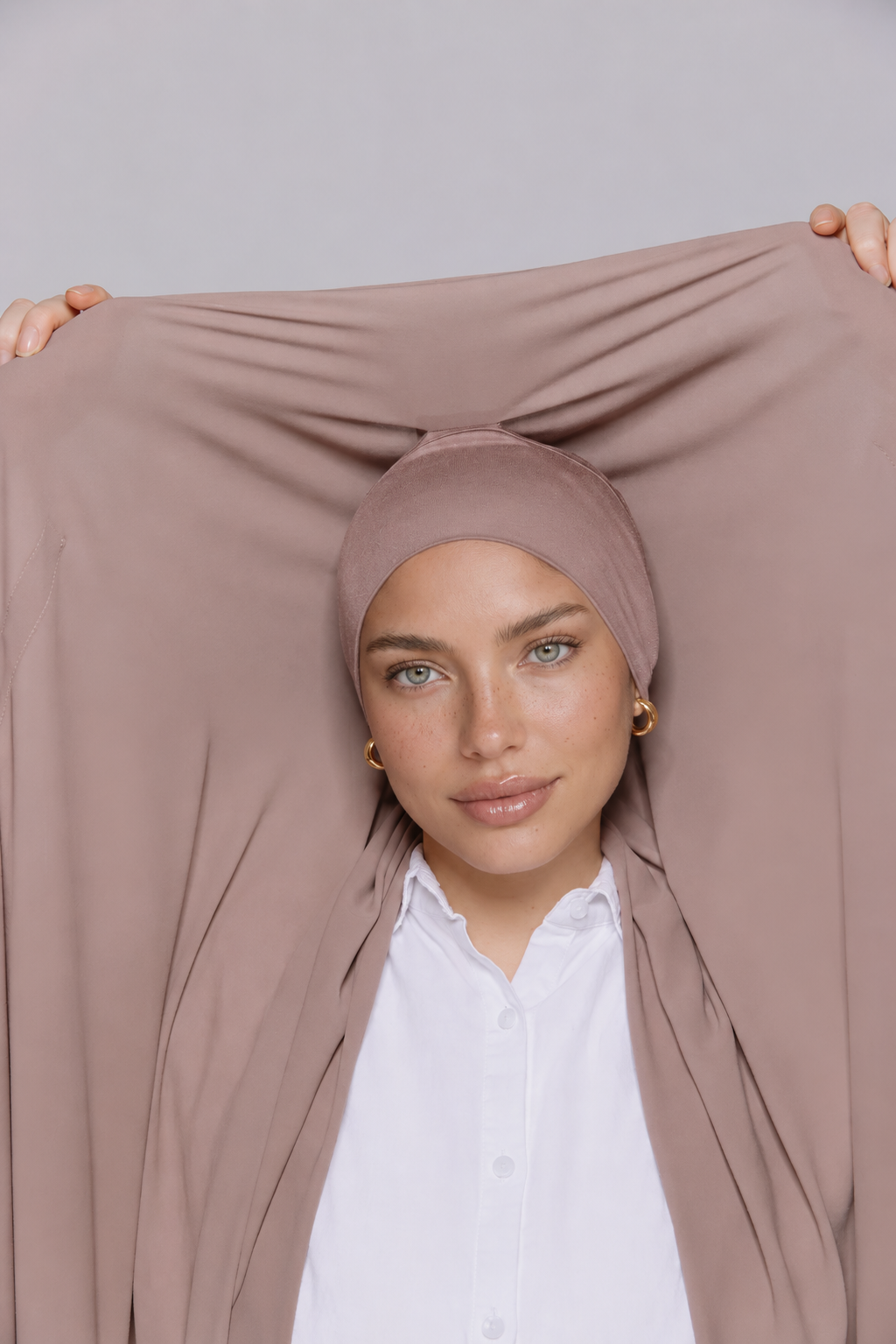A woman wearing a dusty rose headscarf and holding a matching sheer draped fabric panel behind her head, styled over a white collared shirt against a light gray studio background.